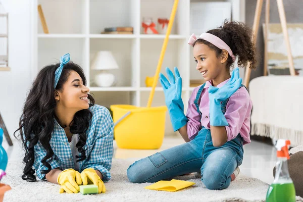 Smiling african american mom and daughter in bright rubber gloves talking while resting on carpet — Stock Photo