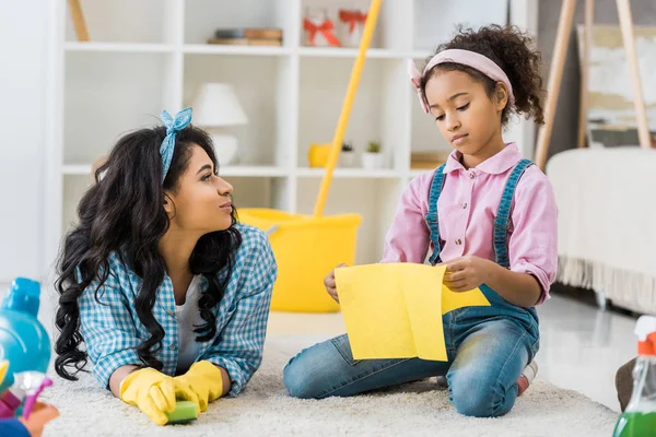 Cute african american child sitting on carpet with yellow rag while mother resting near — Stock Photo