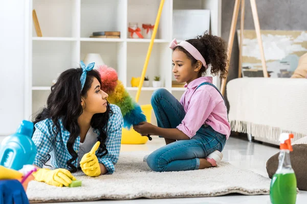 Cute african american child with multicolored duster sitting on carpet while mother lying near — Stock Photo
