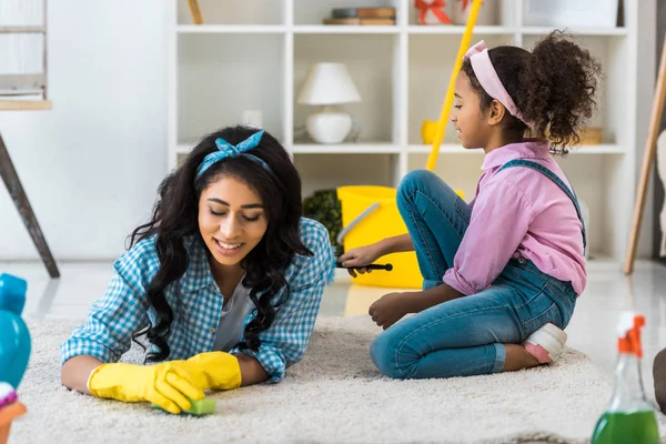Attractive african american woman in yellow rubber gloves lying on carpet while daughter sitting near — Stock Photo