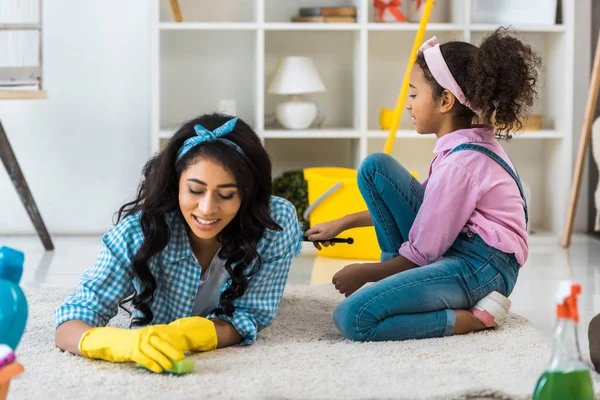 Attraktive Afroamerikanerin in gelben Gummihandschuhen auf Teppich liegend, während Tochter in der Nähe sitzt — Stockfoto