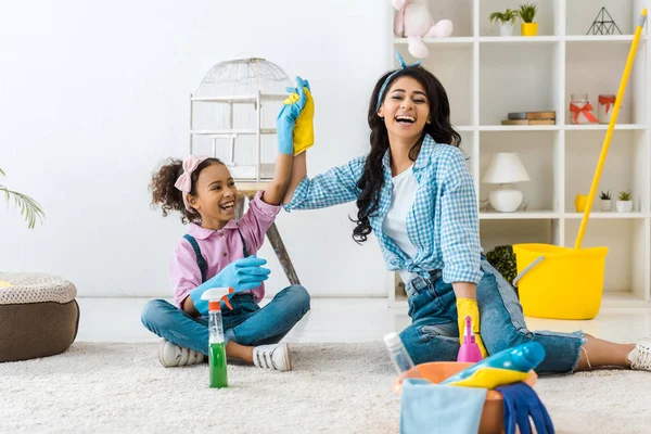 Laughing african american mom holding daughters raised hand while sitting on carpet — Stock Photo