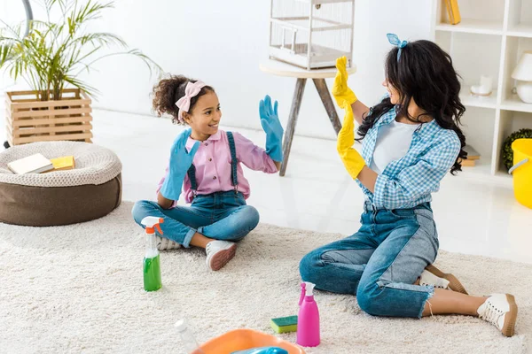 African american mom and cute daughter in bright rubber gloves having fun while sitting on carpet — Stock Photo