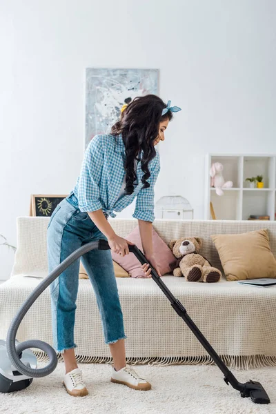 African american woman in plaid shirt and blue jeans vacuuming carpet — Stock Photo