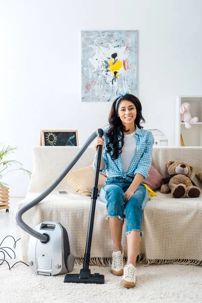 Smiling african american woman sitting on sofa and holding vacuum cleaner rod — Stock Photo