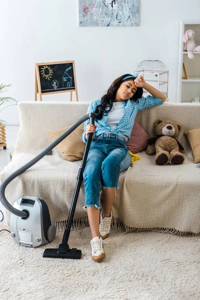 Exhausted african american woman sleeping on sofa while holding vacuum cleaner rod — Stock Photo