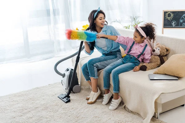 Entzückende afrikanisch-amerikanische Kind sitzt auf dem Sofa mit lächelnder Mutter und hält bunten Staubwedel — Stockfoto