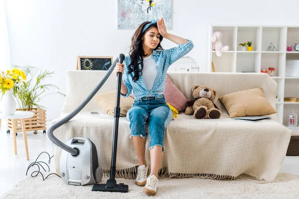 Tired african american woman sitting on sofa while holding vacuum cleaner rod — Stock Photo