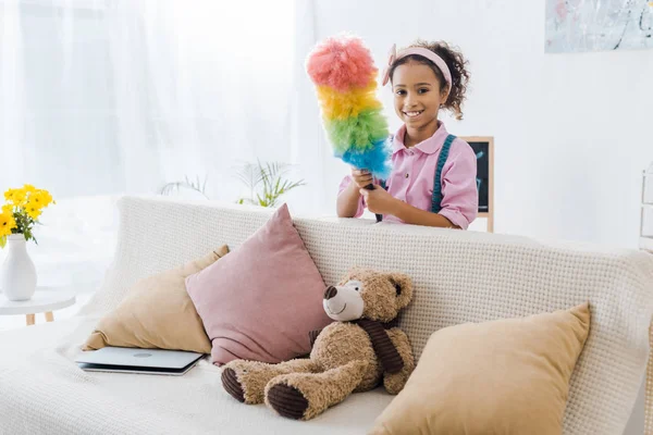 Mignon enfant afro-américain debout près du canapé avec des oreillers et un ours en peluche et tenant un plumeau coloré — Photo de stock