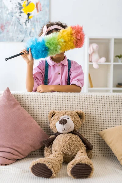Фрикадный американский ребенок прячет лицо с красочным пыльником, стоя рядом с диваном — стоковое фото