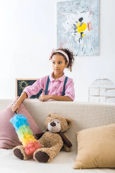 Bonito afro-americano criança espanando sofá com travesseiros e ursinho de pelúcia — Fotografia de Stock