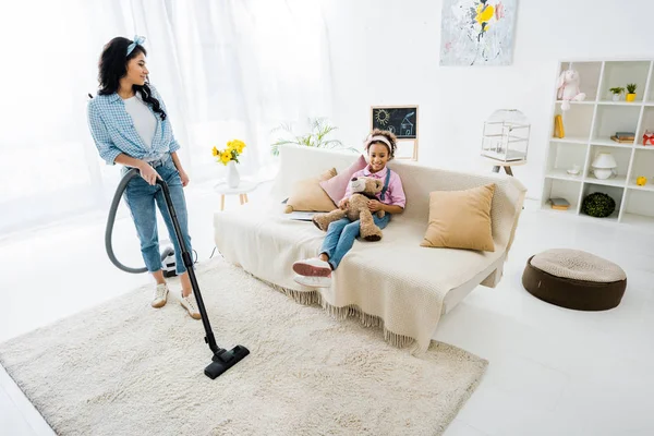 Africano americano madre limpieza alfombra con aspiradora mientras hija sentado en sofá - foto de stock