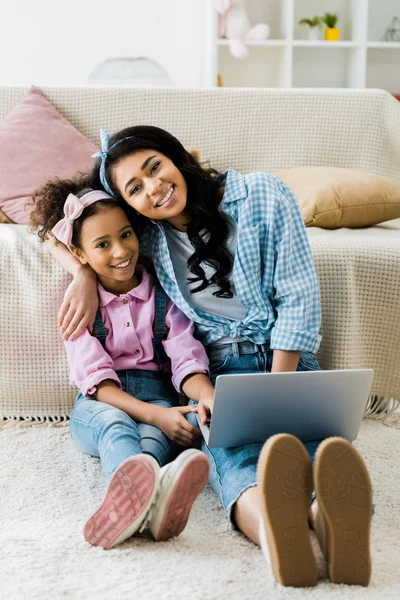 Sonriente afroamericano madre e hija usando el ordenador portátil mientras está sentado en la alfombra cerca del sofá - foto de stock