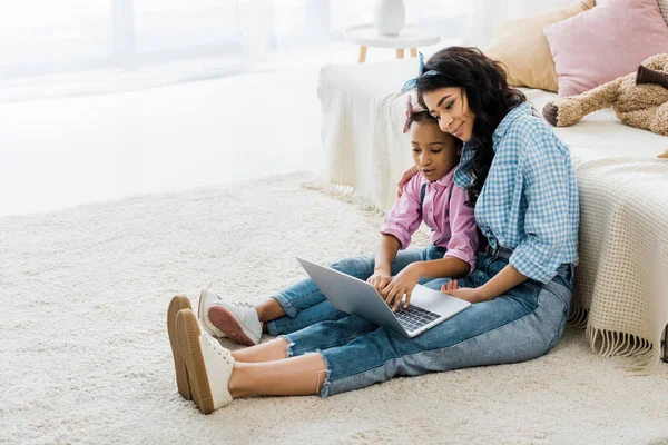 Pretty african american woman with adorable daughter using laptop while sitting on carpet near sofa — Stock Photo