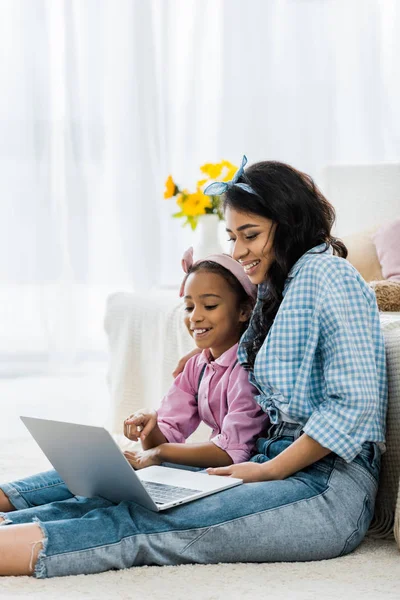 Sorridente madre afroamericana con figlia utilizzando il computer portatile mentre seduto sul tappeto vicino al divano — Foto stock