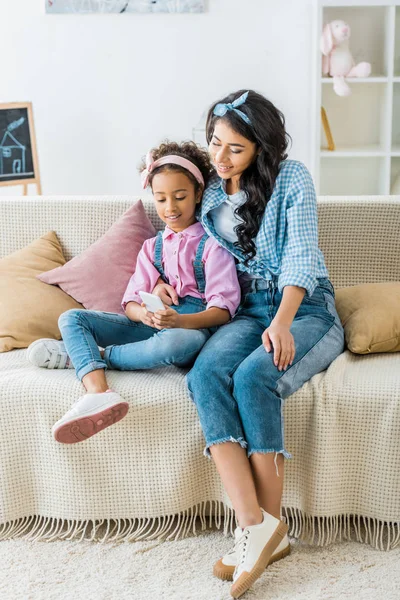 Madre afroamericana y el niño usando el teléfono inteligente juntos mientras están sentados en el sofá en casa - foto de stock