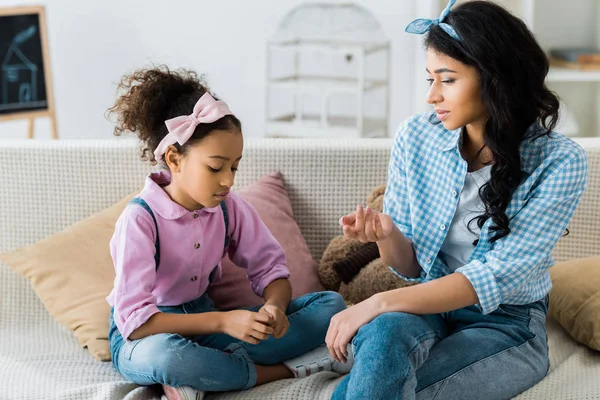 Seria africana americana madre con hija hablando mientras está sentado en sofá - foto de stock