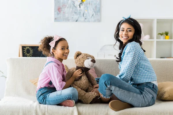 Süße afrikanisch-amerikanische Kind hält Teddybär, während auf dem Sofa mit Mutter sitzen — Stockfoto