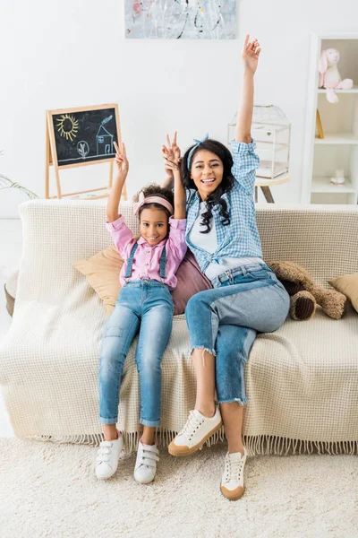 Веселые африканские мама и дочь показывают победные знаки, сидя дома на диване — стоковое фото