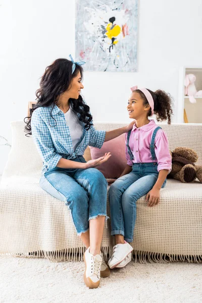 Mujer afroamericana sonriente hablando con su hija mientras está sentada en el sofá en casa - foto de stock