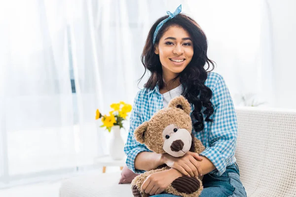 Beautiful african american woman sitting on sofa and holding teddy bear — Stock Photo