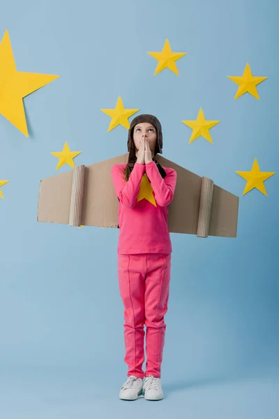 Vista completa de niño con alas de cartón mirando hacia arriba sobre fondo azul estrellado - foto de stock