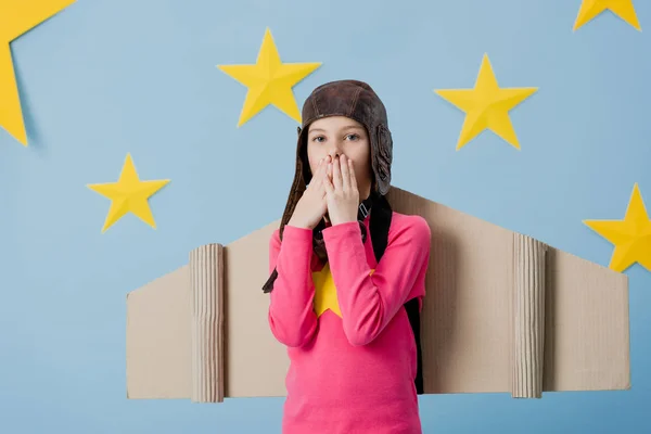 Surprised child with cardboard wings covering mouth with hands on blue starry background — Stock Photo