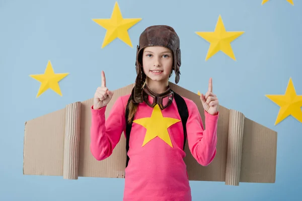 Cute kid with cardboard wings pointing with fingers on blue starry background — Stock Photo