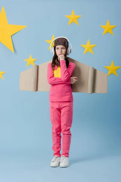 Pensive kid with cardboard wings listening music in headphones on blue starry background — Stock Photo