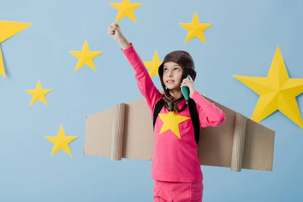 Funny kid with cardboard wings talking on smartphone on blue background with stars — Stock Photo
