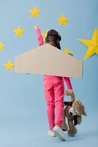 Vista posterior de niño con alas de cartón sosteniendo oso de peluche sobre fondo azul estrellado - foto de stock