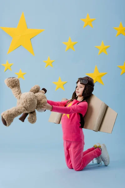 Criança com asas de papelão de pé sobre os joelhos e segurando ursinho de pelúcia no fundo estrelado azul — Fotografia de Stock
