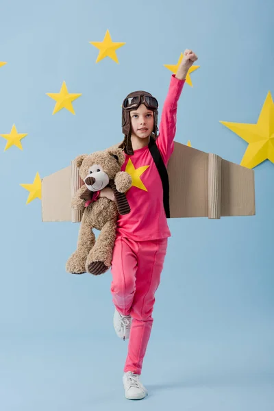 Cute kid with cardboard wings holding teddy bear and posing on blue starry background — Stock Photo