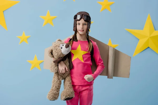 Ребенок с картонными крыльями держит плюшевого мишку на синем фоне со звездами — стоковое фото