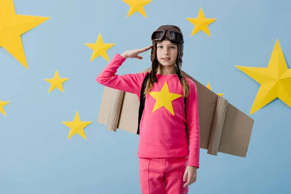 Kid with cardboard wings saluting on blue background with stars — Stock Photo