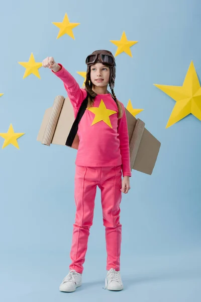 Selbstbewusstes Kind mit Pappflügeln, das die Faust auf blauem Sternenhintergrund hochhält — Stockfoto