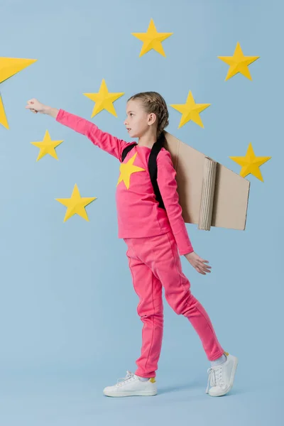 Full length view of kid in pink clothes with cardboard wings holding fist up on blue starry background — Stock Photo