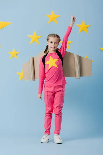 Joyeux enfant avec des ailes en carton debout avec le poing vers le haut sur fond bleu avec des étoiles — Photo de stock