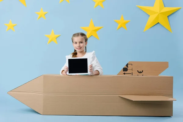 Criança sorridente segurando tablet digital com tela em branco perto de foguete de papelão no fundo estrelado azul — Fotografia de Stock