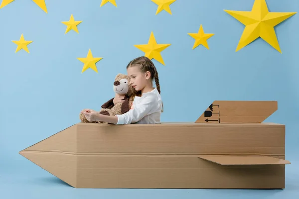 Kid segurando ursinho de pelúcia enquanto brincava com foguete de papelão no fundo estrelado azul — Fotografia de Stock