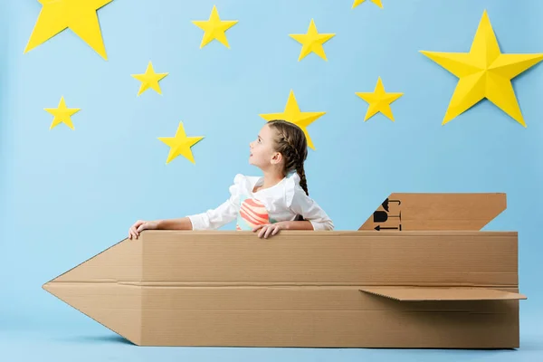 Preteen child sitting in cardboard rocket and looking at stars on blue starry background — Stock Photo