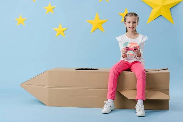 Enfant avec une manette assise sur une fusée en carton sur fond bleu avec des étoiles — Photo de stock