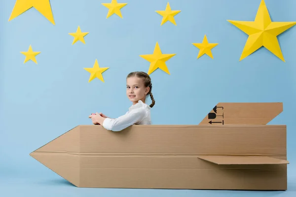 Niño con trenzas sentado en un cohete de cartón y sosteniendo el joystick sobre fondo azul estrellado - foto de stock