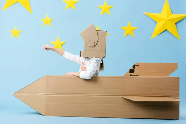 Kid no capacete sentado no foguete de papelão e segurando o punho no fundo estrelado azul — Fotografia de Stock