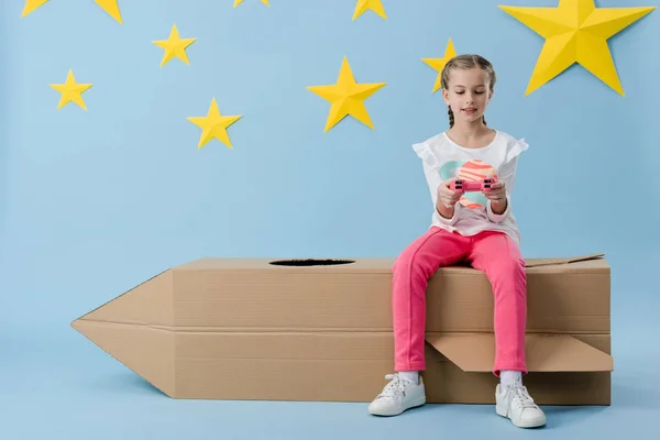 Kid in pink pants sitting on cardboard rocket and holding joystick on blue starry background — Stock Photo