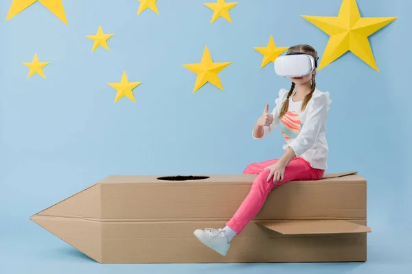 Kid in vr headset sitting on cardboard rocket and showing thumb up on blue starry background — Stock Photo