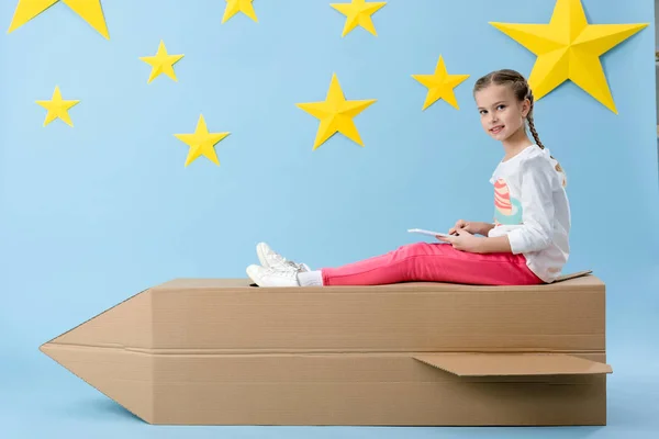 Curious kid with smartphone sitting on cardboard rocket on blue starry background — Stock Photo