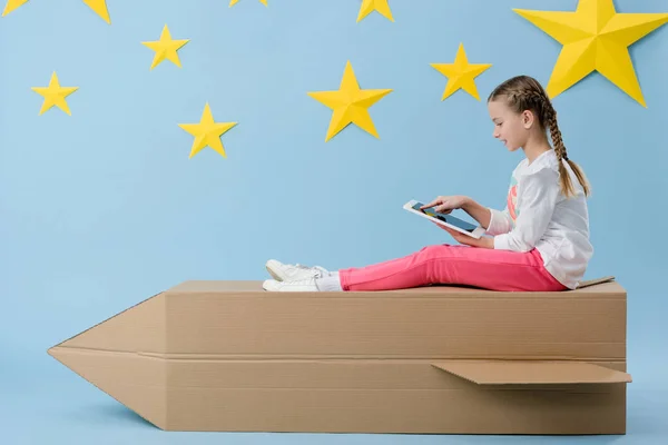 Kind mit Zöpfen sitzt auf Papprakete und nutzt digitales Tablet auf blauem Sternenhintergrund — Stockfoto