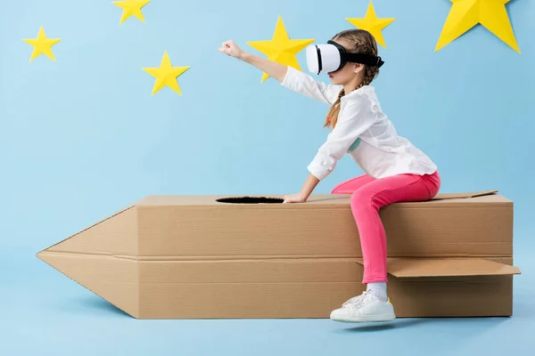 Kid in vr headset sitting on cardboard rocket and holding fist up on blue background with stars — Stock Photo