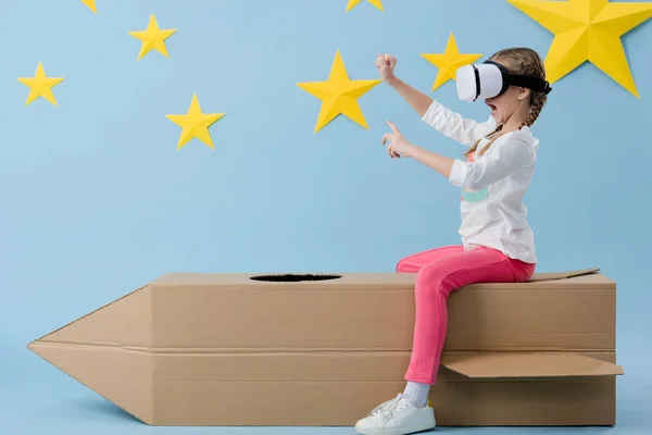 Child in vr headset sitting on cardboard rocket and waving hands on blue starry background — Stock Photo